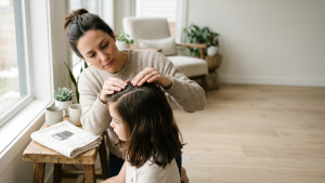 Mother near a bright window gently parting the dark hair of her young daughter at the part line for a careful scalp check, with a fine-toothed metal nit comb resting on a folded white towel on a side table