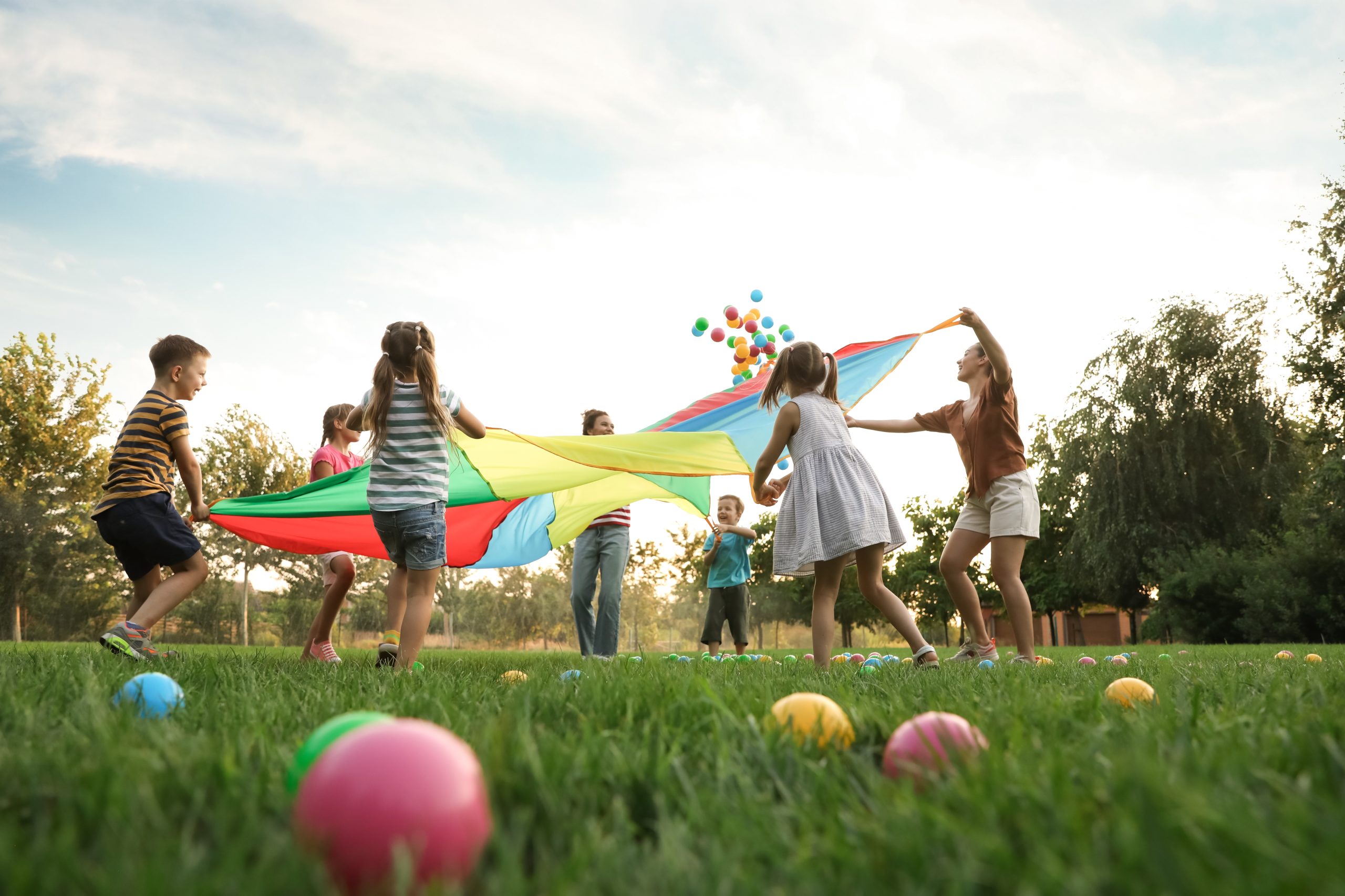 Children and counselors playing with a colorful parachute outdoors at summer camp, a close-contact setting for head lice prevention.
