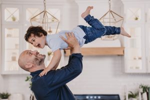 Bald adult dad in a blue shirt lifting his young son in the kitchen, the kind of close household contact moment where lice transfer concerns come up.