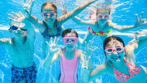 Three young kids swimming underwater in a bright community pool, the kind of summer pool day Palm Beach County parents assume will clear a head-lice case.