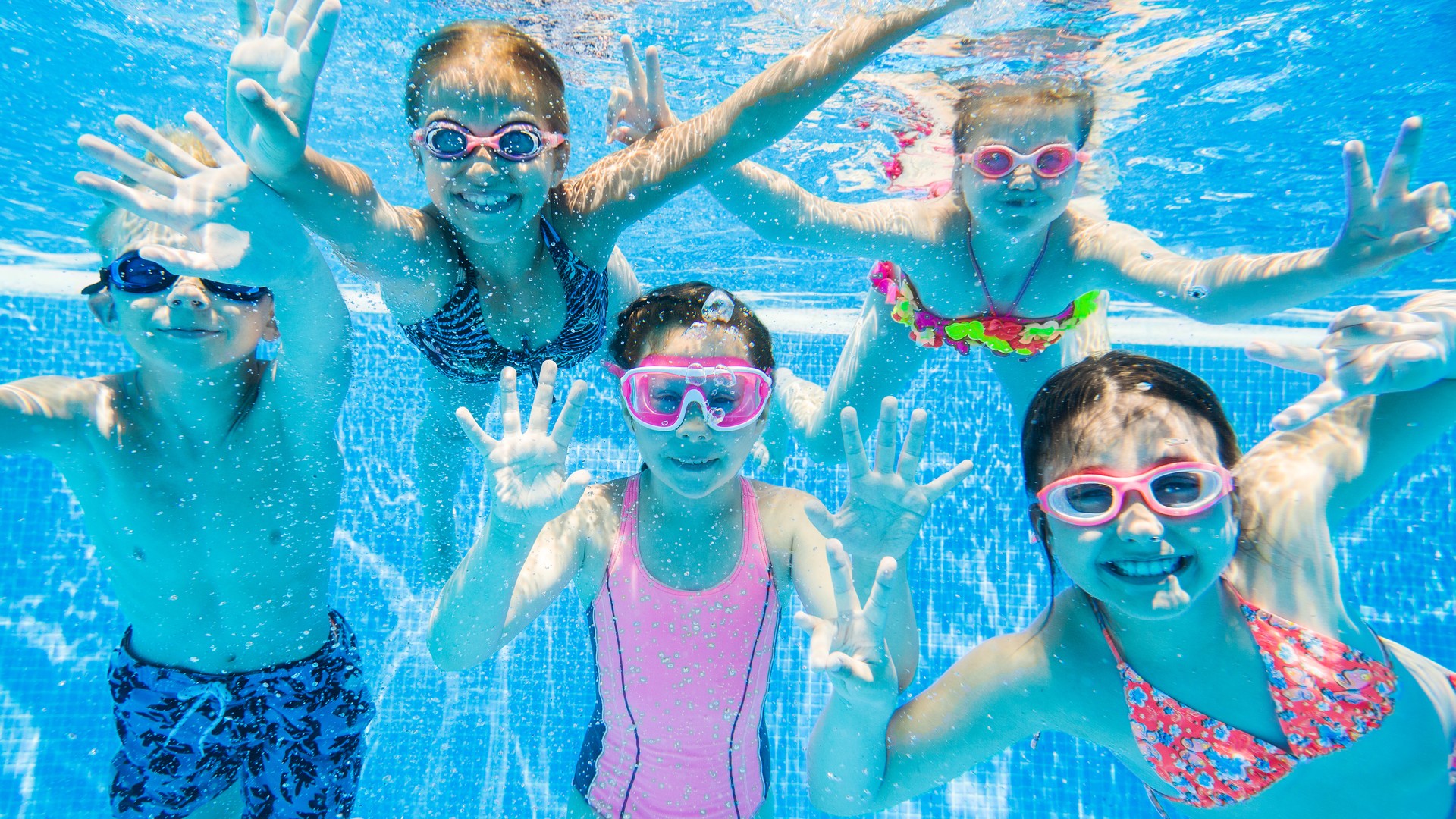 Children swimming underwater in a bright pool, matching parent questions about chlorine, pools, lake water, and head lice.