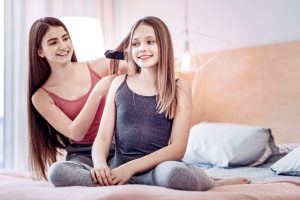 Two young girls in pajamas straightening each other's hair on a bed with a household flat iron, the kind of casual heat-styling moment Palm Beach County parents mistake for a do-it-yourself lice fix.