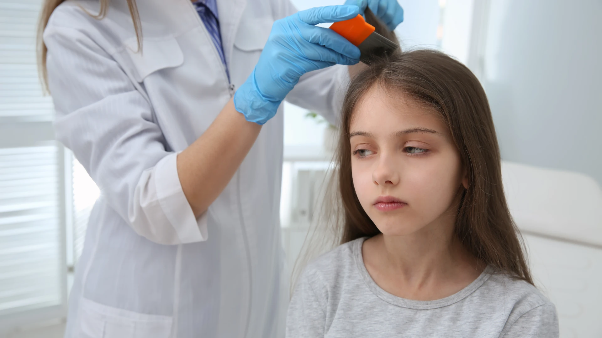 Gloved hands using an orange-handled fine-tooth comb near the crown of a child's brown hair.