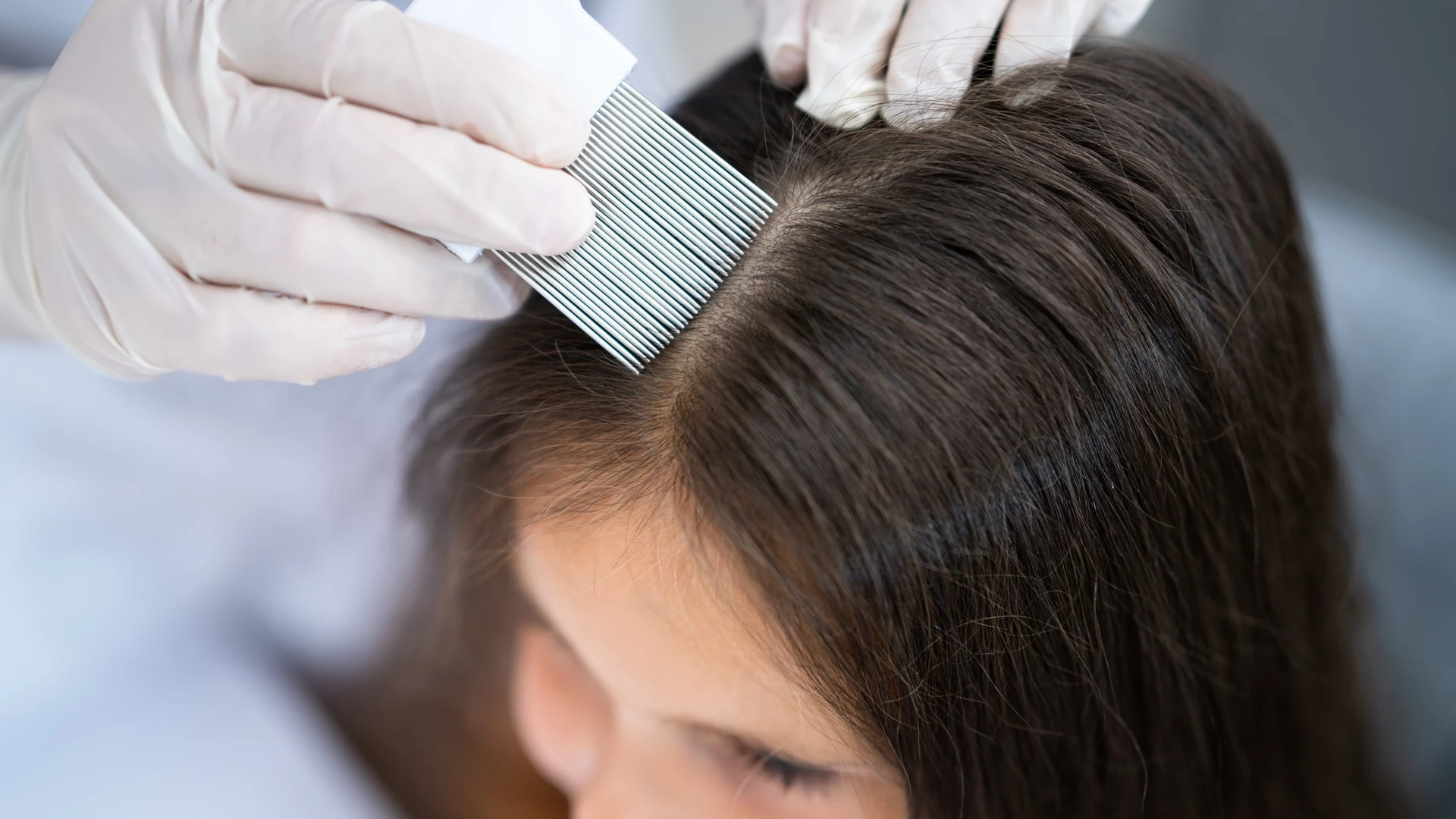 Gloved hands hold a fine-toothed comb in dark hair near the part.